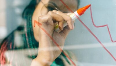 Woman in a Beige Coat Writing on a Glass Panel Using a Whiteboard Marker